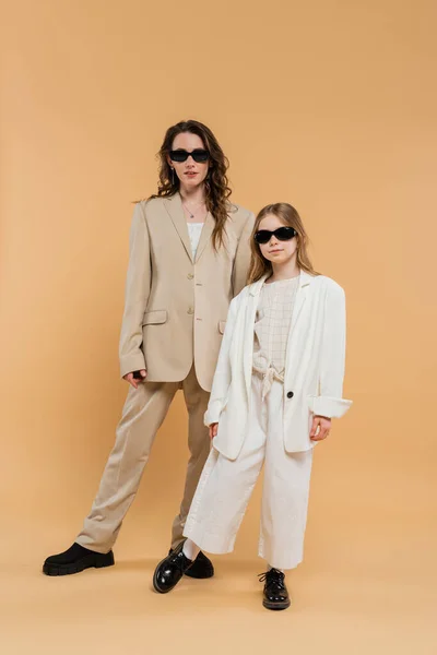 流行の母親と娘でサングラス ビジネス女性と女の子でスーツを着てベージュの背景に一緒に立って ファッショナブルな衣装 正式な服装 企業のお母さん 現代の家族 — ストック写真