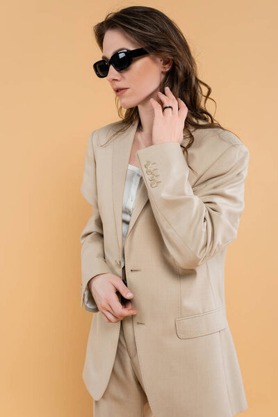 fashion trend concept, young woman with wavy hair standing in fashionable suit and sunglasses on beige background, classic style, chic stylish posing, professional attire, formal attire 