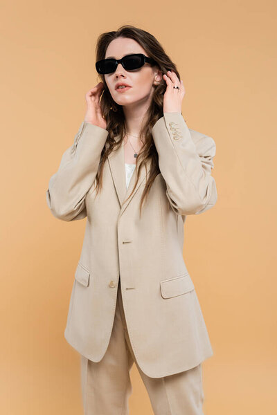 fashion trend concept, young woman adjusting wavy hair and standing in fashionable suit with sunglasses on beige background, classic style, chic stylish posing, professional attire 