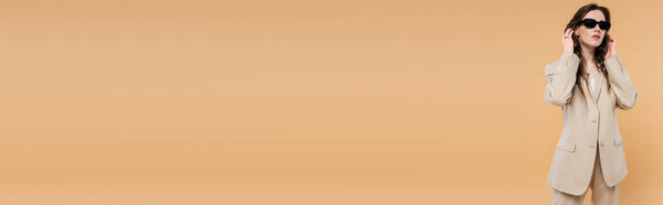 style and fashion concept, young woman adjusting wavy hair and standing in fashionable suit with sunglasses on beige background, classic style, chic stylish posing, professional attire, banner