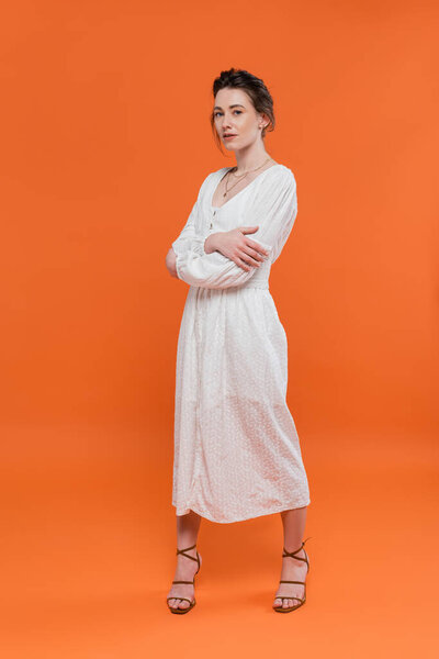 летние тенденции, модель в белом солнцезащитном платье глядя на камеру и стоя со сложенными руками на оранжевом фоне, стильная поза, дама в белом, яркий фон, мода, молодая женщина 