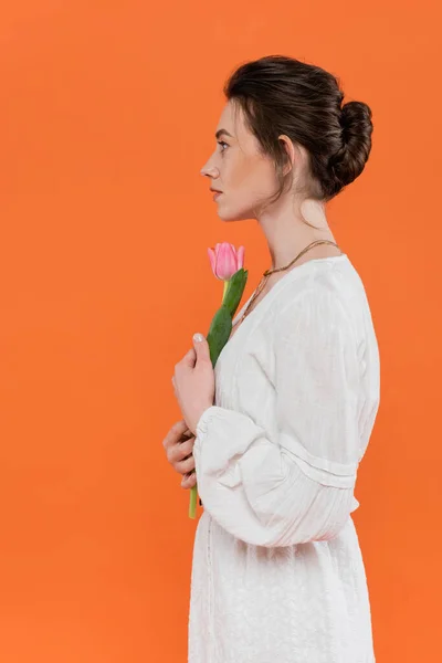 stock image side view of young woman in white sun dress holding pink tulip and standing on orange background, lady in white, vibrant background, fashion, summer, portrait, sensuality, everyday fashion 