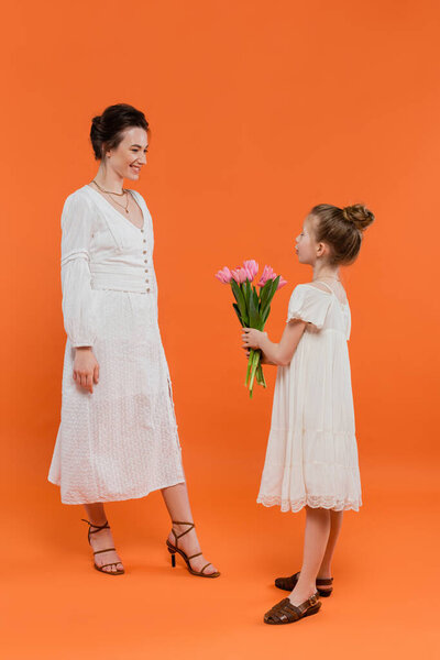 День матери, милая девочка-подросток держа букет цветов рядом с матерью на оранжевом фоне, связи, белые платья, розовые тюльпаны, счастливый праздник, яркие цвета, радостное событие 