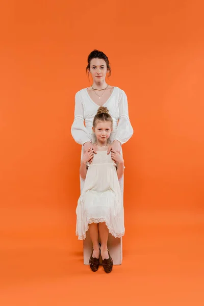 stock image motherly love, young mother hugging daughter and standing together in white sun dresses on orange background, family fashion, fashionable woman and girl, female bonding, modern parenting 