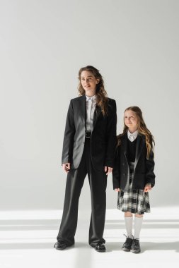 Çalışan anne ve okul kızı, okul üniformalı neşeli kız gri arka planda iş kadınıyla birlikte duruyor, el ele tutuşuyor, resmi giyiniyor, moda ailesi, bağ kuruyor, modern ebeveynlik yapıyor. 