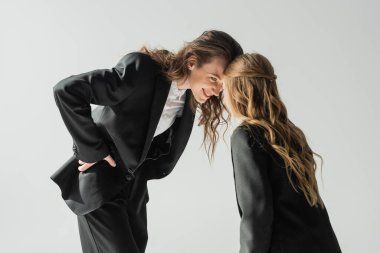 happy mother and daughter having fun, woman in suit and schoolgirl in uniform looking at each other and smiling on grey background in studio, fashionable family, formal attire clipart