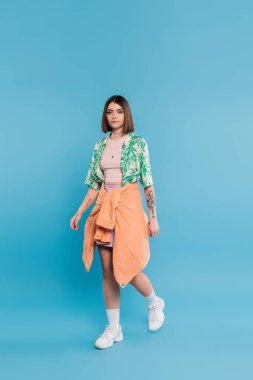 günlük stil, kısa saçlı genç esmer kadın palmiye ağacı desenli gömlek, etek ve beyaz spor ayakkabılı mavi arka planda, tasasız, dövmeli, burun piercingli, günlük kıyafet, tam boy 