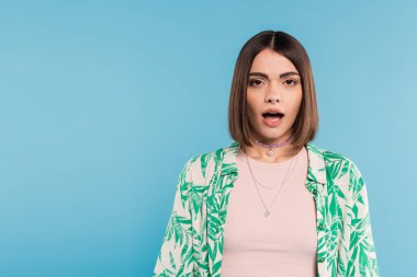 Ağzı açık duygusal bir kadın, şok olmuş bir model mavi arka planda kameraya bakıyor, palmiye ağacı desenli gömlek, burun piercingli, günlük kıyafet, Z jenerasyonu, şaşırmış yüz. 