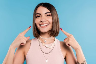 Heyecan, dövmeli kısa esmer saçlı, mavi arka planda gülümseyen ve yanaklarını gösteren genç bir kadın, günlük kıyafetler, genel z moda, mutluluk, neşe dolu 