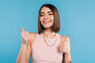 Pozitiflik, kısa esmer saçlı, kısa saçlı, mavi arka planda gülümseyen ve başparmaklarını gösteren genç bir kadın, günlük kıyafetler, genel z moda, mutluluk, jest gibi.