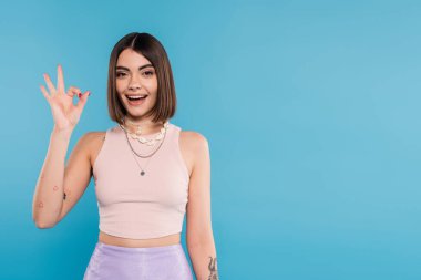 Heyecan, kısa saçlı, kısa saçlı kısa saçlı, gülümseyen ve mavi arka planda güzel jestler yapan, günlük kıyafetler, moda, moda, mutluluk gösteren genç bir kadın. 