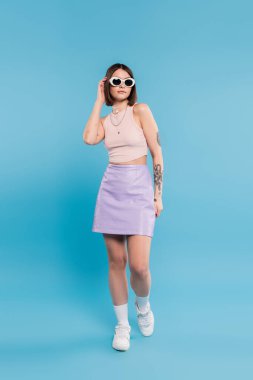 Moda trendi, kısa saçlı, atlet bluzlu, etekli, beyaz spor ayakkabılı, güneş gözlüklü, mavi arka planda poz veren esmer genç kadın, günlük kıyafetler, genel z moda, kişisel tarz, burun piercing 'i. 