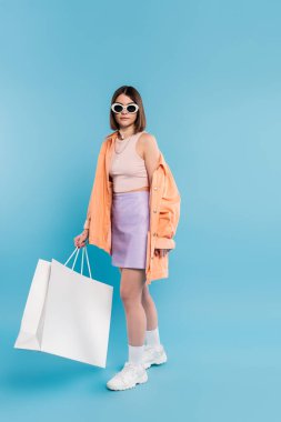 Yaz alışverişi, kolsuz bluz, etek, güneş gözlüğü ve turuncu gömlekli esmer kadın mavi arka planda alışveriş çantasıyla poz veriyor, günlük giyiniyor, şık poz veriyor. 