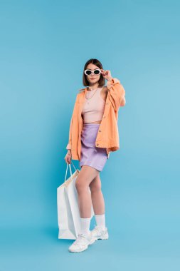 Moda alışverişi, kolsuz bluz, etek, güneş gözlüğü ve turuncu gömlekli esmer kadın mavi arka planda alışveriş çantasıyla poz veriyor, günlük kıyafet, şık poz, genel moda z, modern moda. 