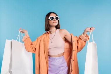 Yaz cümbüşü, güneş gözlüklü esmer bir kadın mavi arka planda alışveriş çantalarıyla poz veren modaya uygun giyinen, şık poz veren, z veren, modern moda modası olan Z kuşağı. 