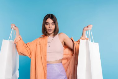 yaz trendleri, dekolteli bir kadın mavi arka planda alışveriş çantalarıyla poz veriyor, günlük kıyafetler, tarz, z nesli, modern moda, turuncu gömlek, kolsuz bluz ve etek 