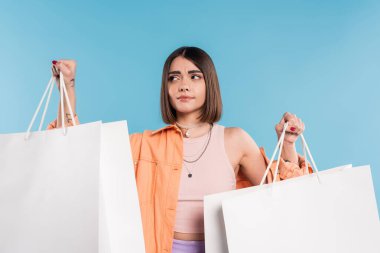 Alışveriş çılgınlığı, moda kıyafetleri içinde kafası karışık genç bir kadın mavi arka planda alışveriş çantalarıyla poz veriyor, günlük kıyafetler, şık, Z jenerasyonu, modern moda, yaz kıyafetleri 