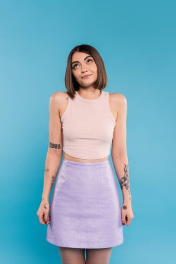 not knowing, smiling young woman with tattoos and nose piercing standing in tank top and skirt on blue background, looking up, confused, pretty face, generation z, summer outfit  clipart