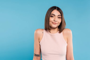 not knowing, smiling young woman with tattoos and nose piercing standing in tank top on blue background, looking at camera, confused, pretty face, generation z, summer outfit  clipart