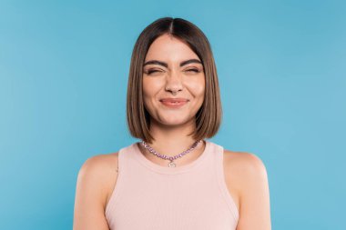 Pozitif, kısa saçlı, burun piercingli, mavi arka planda gülümseyen neşeli genç bir kadın. Z nesli, modaya uygun, kişisel tarzda, gümüş kolye, duygusal, mutluluk.  