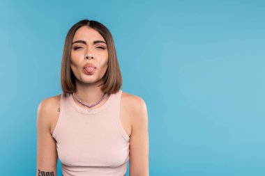 Dövmeli, burun piercingli, kısa saçlı, mavi arka planda dili olan genç bir kadın. Z nesli, modaya uygun bir model, moda yaz modası, gençlik kültürü.  