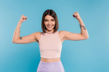 women power, cheerful young woman with short hair, tattoos and nose piercing showing muscles on blue background, generation z, displeased, casual attire, strength  clipart