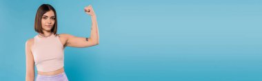 female power, brunette young woman with short hair, tattoos and nose piercing showing muscle on blue background, generation z, displeased, casual attire, strength, banner  clipart
