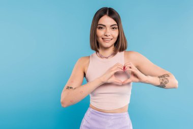 positivity and love, happy young woman with short hair, tattoos and nose piercing showing heart gesture with hands on blue background, generation z, cheerful, casual attire clipart