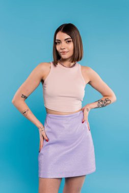 Yazlık kıyafet, günlük kıyafet, kısa saçlı, dövmeli ve burnu piercingli mutlu genç kadın elleri mavi arka planda, Z neslinin günlük tarzında ayakta duruyor.