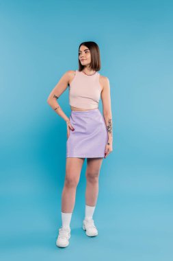 Yaz trendleri, kısa saçlı, atlet bluzlu, etekli, beyaz spor ayakkabılı, mavi arka planda poz veren esmer bir kadın, günlük kıyafet, genel z moda, kişisel tarz, burun piercing, el kalçada. 