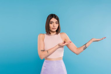Kısa saçlı esmer bir kadın mavi arka planda bir şey gösteriyor, günlük kıyafet, genel z moda, kişisel tarz, burun piercing, elleriyle işaret ediyor. 