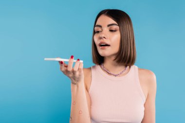 sesli mesaj, kısa saçlı, dövmeli ve burun piercingli mavi arka planda akıllı telefon kullanan genç bir kadın, günlük kıyafet, genel z moda, kişisel tarz, sosyal medya etkileyicileri 