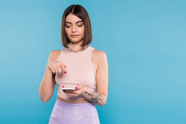 Kısa saçlı, kısa saçlı, burnu delinmiş, mavi arka planda cep telefonu kullanan, günlük kıyafet giyen, genel moda, sosyal medya etkisinde olan genç bir kadın. 