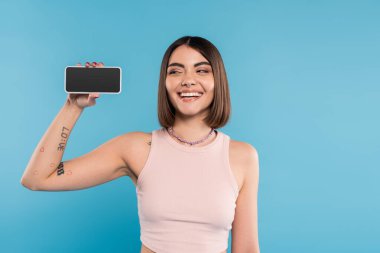 Akıllı telefon, beyaz ekran, kısa saçlı, dövmeli ve burun piercingli mutlu genç kadın cep telefonunu mavi arka planda tutuyor, genel z modası, sosyal medya etkileri 