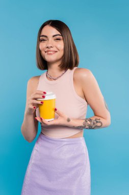 Kahve içmek için, kısa saçlı, dövmeli ve burnu piercing 'li mutlu genç bir kadın elinde mavi arka planda kağıt bardak, z nesli, yaz trendi, çekici, kahve kültürü, günlük tarz 