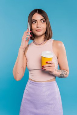 Kahveye gitmek, mutlu ve dövmeli kısa saçlı ve burunlu genç bir kadın elinde kağıt bardakla piercing takıyor ve mavi arka planda akıllı telefondan konuşuyor. Z nesli, yaz trendi, çekici, kahve kültürü. 