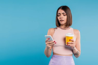 Kahve, sosyal medya etkileri, kısa saçlı ve burunlu dövmeli genç bir kadın elinde kağıt bardakla geziyor ve mavi arka planda akıllı telefon kullanıyor, Z nesli, yaz trendi 