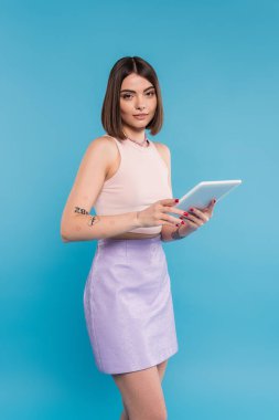 Esmer, kısa saçlı, dövmeli ve burun delici dijital tablet dizüstü bilgisayarı mavi arka planda, z nesli, yaz eğilimleri, çekici, sosyal medya etkileri, tablet kullanıcısı 