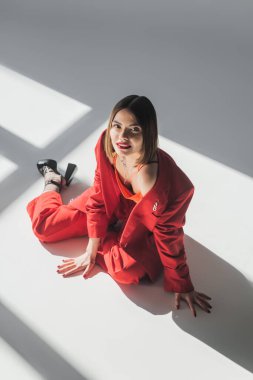Üst tabaka, kısa saçlı, gri arka planda oturan esmer bir kadın, z jenerasyonu, modaya uygun kıyafetler, profesyonel kıyafetler, yönetici tarzı, topuklu ayakkabılar, kırmızı giyen bayan. 
