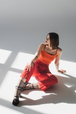 Uzun boylu, kısa saçlı, takım elbiseli, gri arka planda oturan genç bir kadın, z nesli, modaya uygun bir model, profesyonel kıyafet, kurumsal moda, topuklu ayakkabı, kırmızı elbiseli bir bayan. 