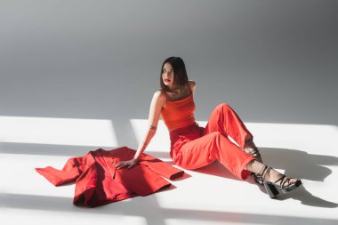 Uzun boylu, kısa saçlı bir kadın gri arka planda kırmızı ceketin yanında oturuyor. Z nesli, modaya uygun bir model, profesyonel kıyafet, kurumsal moda, topuklu ayakkabı, kırmızı giyen bayan. 