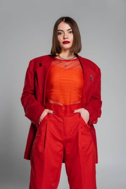 Moda görünüşü, kısa saçlı ve burun piercingli genç bir kadın gri arka planda poz veriyor, Z jenerasyonu, modaya uygun kıyafetler, profesyonel kıyafetler, cepte eller, kalın makyaj. 