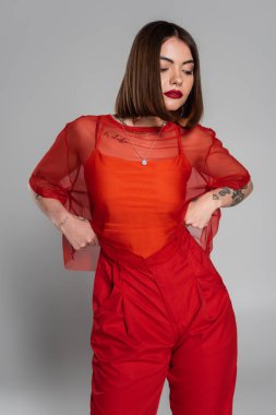 Şık kırmızı elbise, dövmeli ve kısa saçlı ve burnu piercingli esmer kadın şeffaf bluz ve pantolonla gri arka planda poz veriyor, modern tarz, z jenerasyonu, moda
