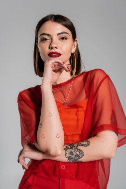 moda görünüş, kırmızı kıyafet, dövmeli ve kısa saçlı esmer kadın ve burun piercing gri arka planda şeffaf bluz poz, modern tarz, z nesil z, portre 
