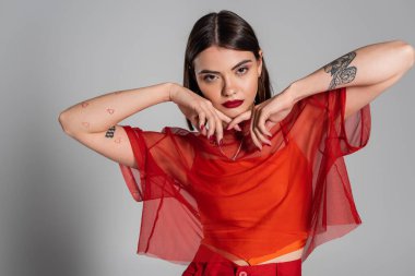 Z nesli, dövmeli ve esmer genç kadın portresi kısa saçlı ve burun piercingli gri arka planda şeffaf bluz giymiş, modern tarzda, moda görünümlü, kırmızı elbiseli.