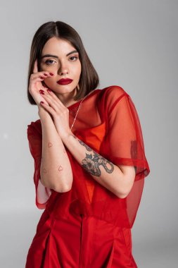 Model, kırmızı elbise, kısa saçlı ve burun piercingli genç kadın gri arka planda kırmızı şeffaf bluzla poz veriyor, modern tarz, z nesli, moda
