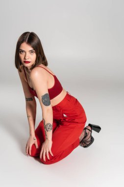 Kırmızı elbiseli genç manken, kısa saçlı ve burnu piercingli esmer kadın gri arka planda kırmızı bluz ve pantolonla poz veriyor, modern tarz, z jenerasyonu, moda trendi. 