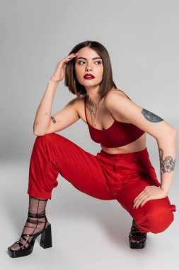 Moda trendi, kırmızı elbiseli genç manken, kısa saçlı ve burnu piercing 'li kadın gri arka plan, modern tarz, tam boy z üzerinde otururken kırmızı hasat bluzu ve pantolonuyla poz veriyor. 