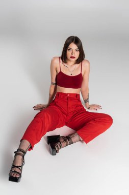 Moda trendi, kırmızı elbiseli genç bir model, kısa saçlı ve burnu piercingli bir kadın gri arka planda kırmızı bluz ve pantolonla poz veriyor, Z jenerasyonu, tam boy, yüksek açı manzaralı. 