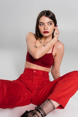 Moda modası, şık bir tarz, kırmızı elbiseli genç bir model, burnu piercing 'li kırmızı bluzla poz veren ve gri arka planda pantolonu olan, z nesli, tişörtünü ayarlayan kadın.